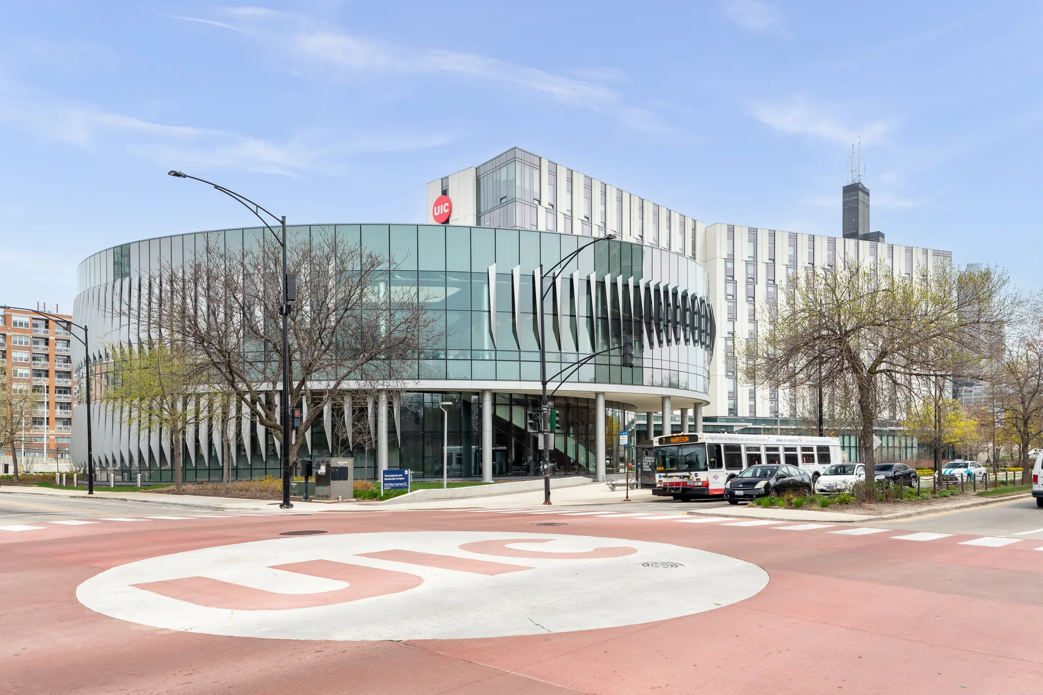 Academic and Residential Complex at the University of Illinois Chicago in Chicago, IL. Modern circular glass-and-white UIC medical center building on a city block with a large painted UIC logo in the foreground of the intersection, buses and cars at the curb, leafless trees and the Chicago skyline behind — portrait of Dr. Ali Dalal’s UIC-affiliated clinical site.