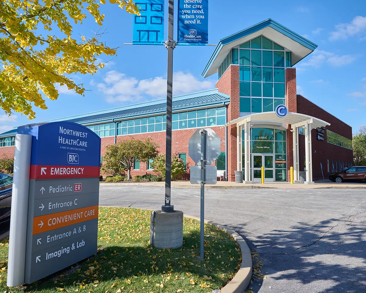Northwest Health Care Northwest HealthCare brick outpatient clinic building with teal metal roof and glass-front entry labeled Entrance C, foreground directional sign listing Emergency, Pediatric ER, Convenient Care, Entrance A & B, and Imaging & Lab, landscaped parking lot with autumn leaves and a blue BJC HealthCare banner on a light pole