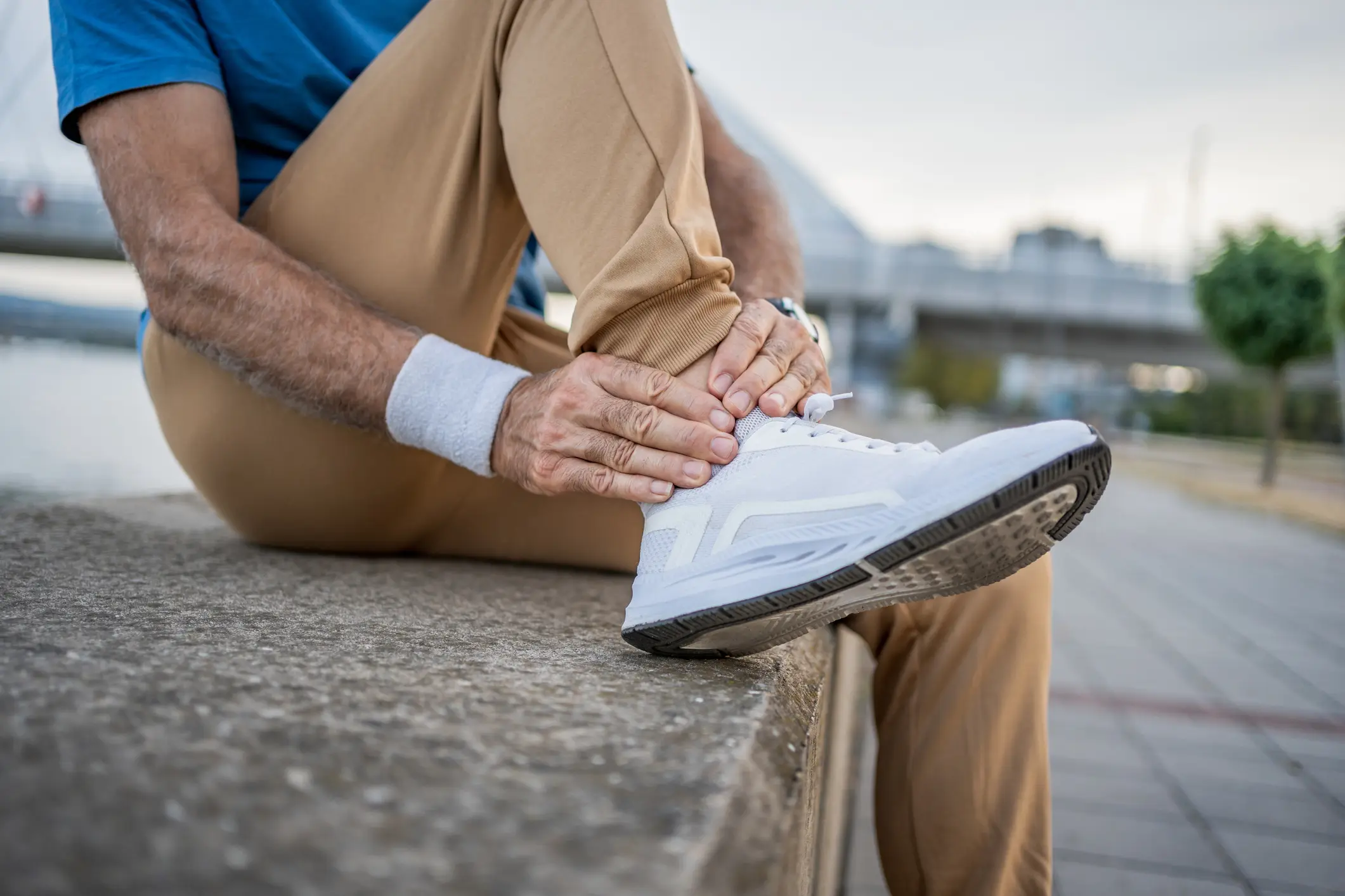 Man in casual athletic wear seated on a riverside ledge holding his lower right leg and ankle, white sneaker visible, suggesting ankle or foot pain and need for orthopedic care.