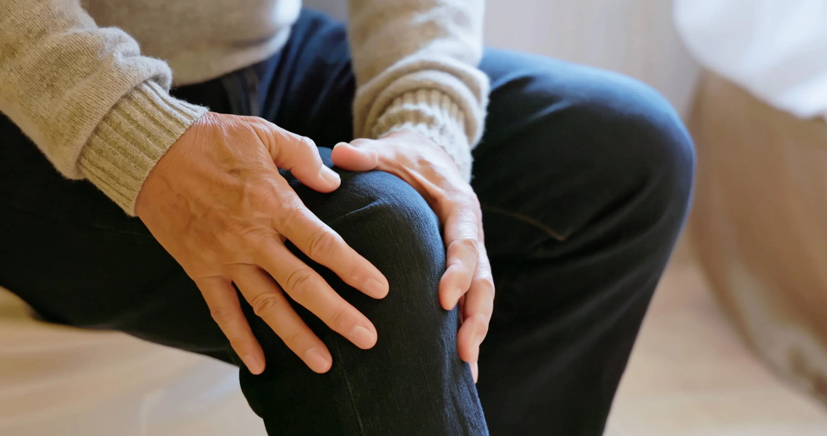 Elderly person sitting on a couch with both hands gently holding a painful right knee, illustrating knee pain and orthopedic care for hip, knee and ankle conditions
